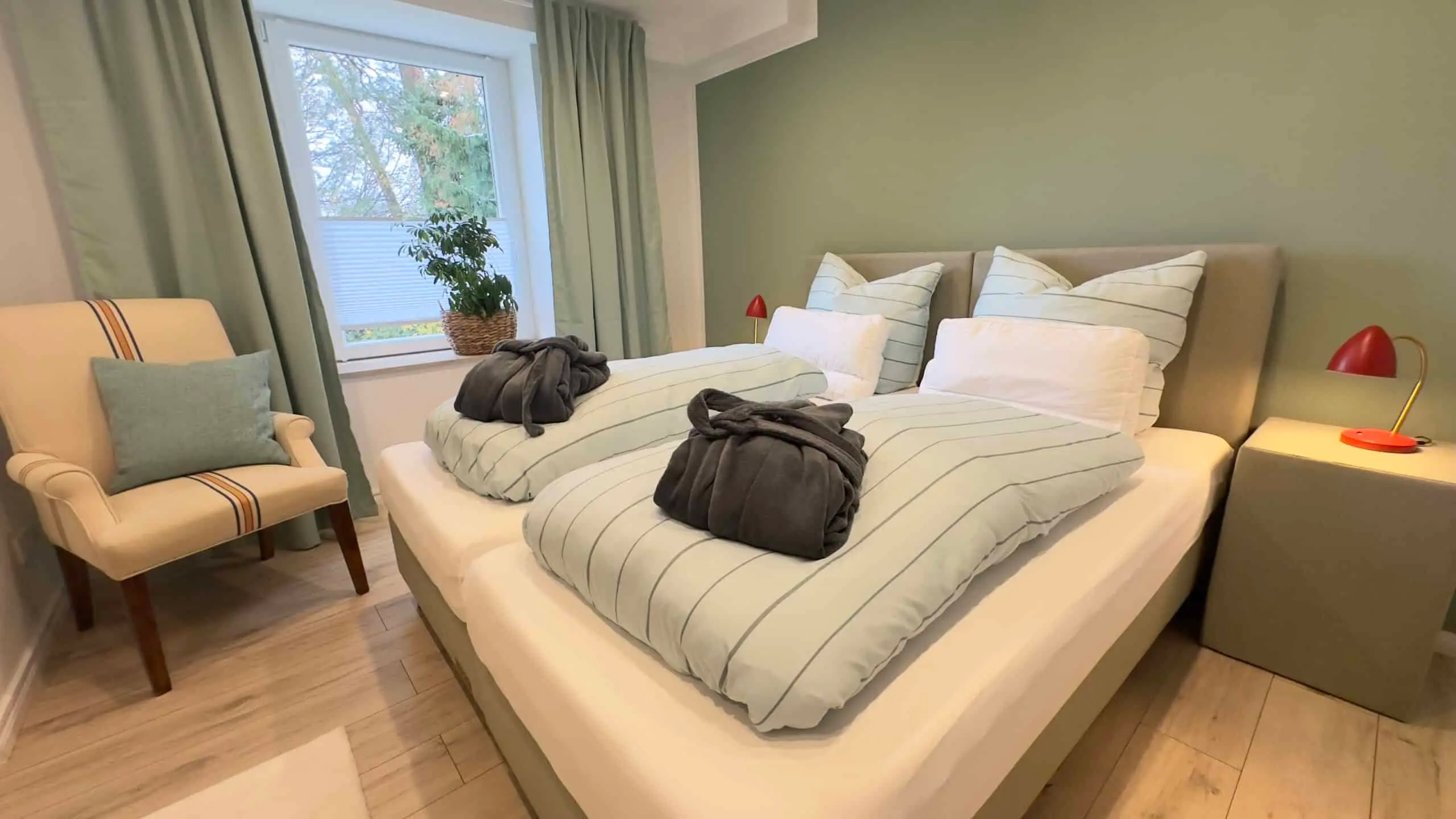 Schlafzimmer der Ferienwohnung „Zur Gelassenheit“ mit salbeigrüner Wand, Boxspringbett und Sessel am Fenster
